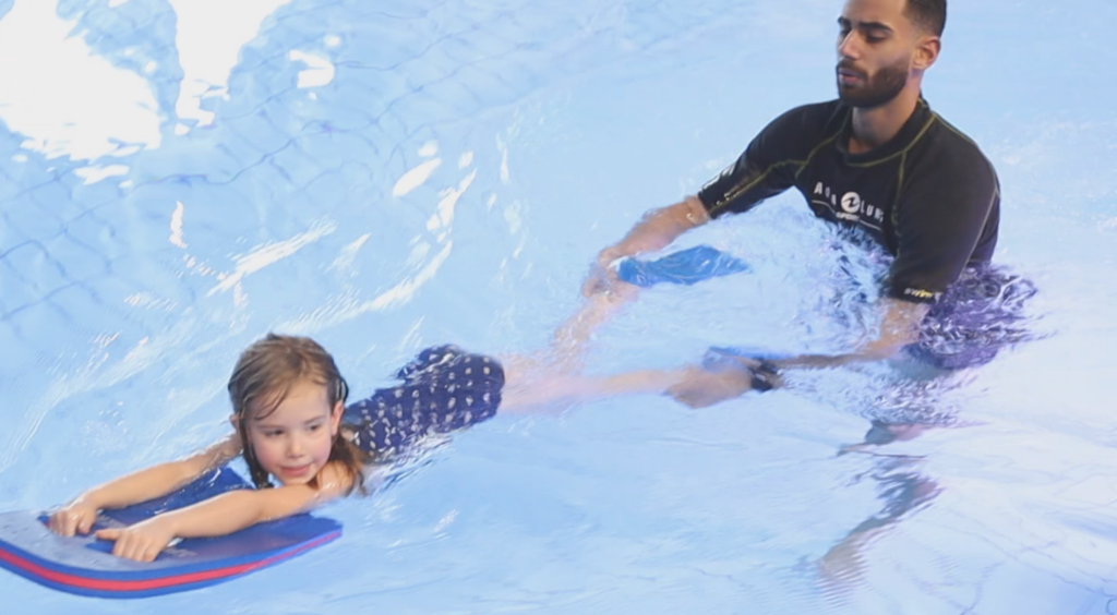 Kind und Arman Moradi üben die Strampelbeine. Das Kind schwimmt mit Schwimmbrett und Flossen und Arman Moradi hält die Knöchel des Kindes.