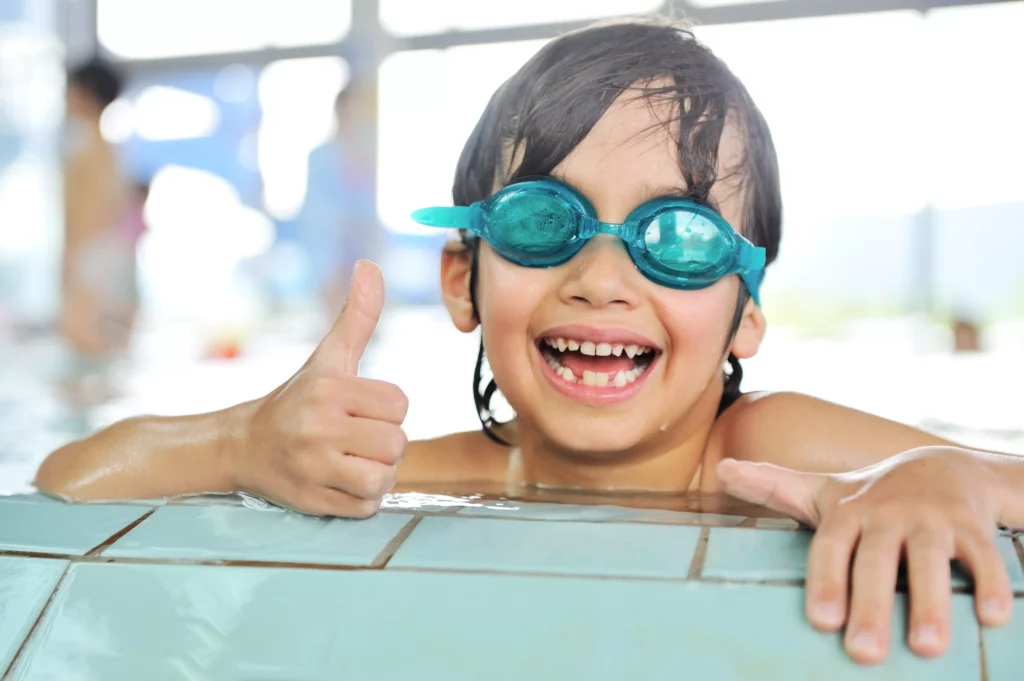 Kind im Schwimmbad zeigt einen Daumen nach oben und lacht, weil er das Schwimmen liebt.
